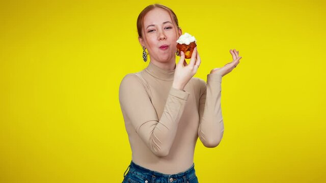 Happy young woman eating sweet delicious muffin at yellow background. Portrait of smiling joyful Caucasian lady enjoying tasty snack. Confectionery concept.