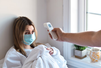 Young teenage girl laying in bed sick and having her temperature taken by non contact thermometer 