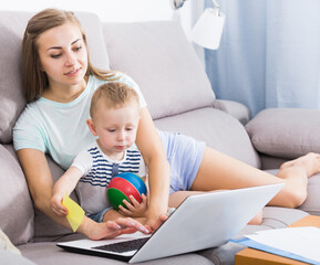 Satisfied mother with child is productively working behind laptop at home.