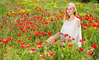 Beautiful woman wearing white dress sitting in poppy field and enjoying summer day