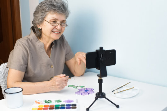 Smiling senior woman taking painting course online. An elderly woman in glasses paints flowers with watercolors, indoors. Home distance learning, retirement hobby concept