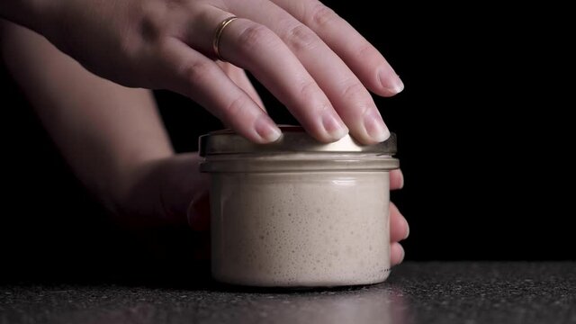 Female hands opening a jar of live leaven for wheat sourdough bread in slow motion against a black background with copy space. High quality FullHD footage