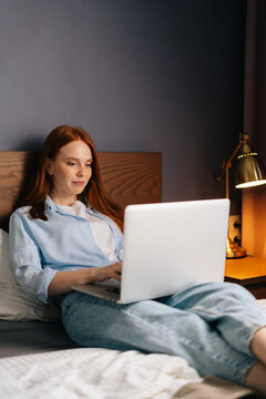 Cute redhead young woman lying on bed and using computer laptop for using social media. Concept of leisure activity red-haired female at home during self-isolation.