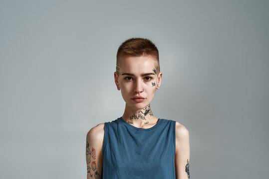 Portrait of tattooed young caucasian woman with piercing looking at camera while posing isolated over gray background