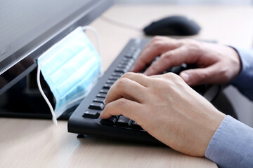 Male hands on PC keyboard and removed medical mask on a desk. Man at computer sitting near the...
