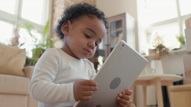 Low angle close up of adorable black toddler boy sitting on floor in living room and looking at tablet