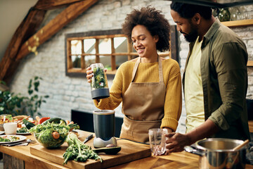 Happy black couple blending vegetables while making smoothie in the kitchen.