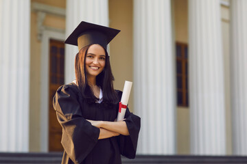 Portrait of confident female student with diploma in hands on university background. Girl is...