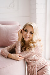 Naklejka premium Attractive young blonde woman with curly hair and makeup in a sequined dress in the bright interior of the studio house