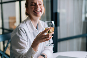 Close-up portrait of laughing redhead young woman holding glass with champagne near face, looking away. Portrait of pretty female with glass of wine.