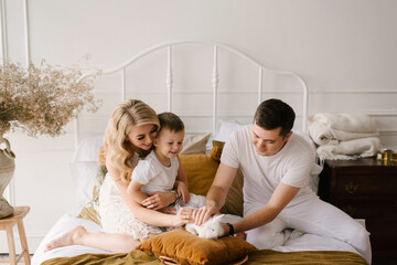 Beautiful young family man woman and son in white clothes play on the bed with a rabbit at home