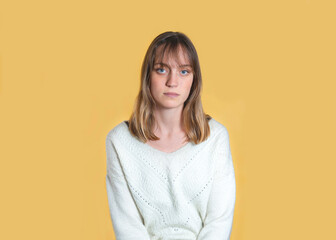 Studio portrait of a woman. She has blond hair, a fringe and blue eyes. She looks serious. She is standing in the center of the image, in front of a yellow background.