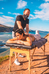A young man performing a massage in nature on the coast near the sea, masseuse with a face mask in the coronavirus pandemic