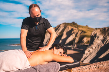 Relaxed girl in the massage on the coast near the sea, masseuse with face mask in the coronavirus...