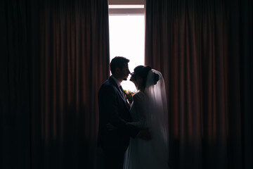 Beautiful and loving honeymoon hut at the window with curtains in the hotel room. Wedding portrait of stylish groom wearing suit and cute bride with curly hair.
