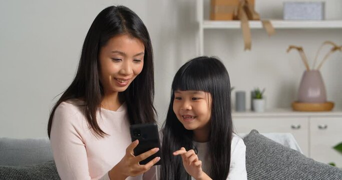 Calm focused asian woman young single mother nanny older sister sitting on sofa with little girl daughter child looking at screen of mobile smartphone ordering shopping online using app online service
