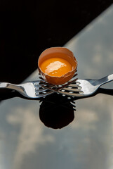Broken fresh chicken egg (Hen egg) balanced on a composition of two intertwined forks over black floor. Copy space, Selective focus.
