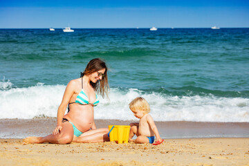 Pregnant mother and little toddler boy play together near sea waves on summer vacation