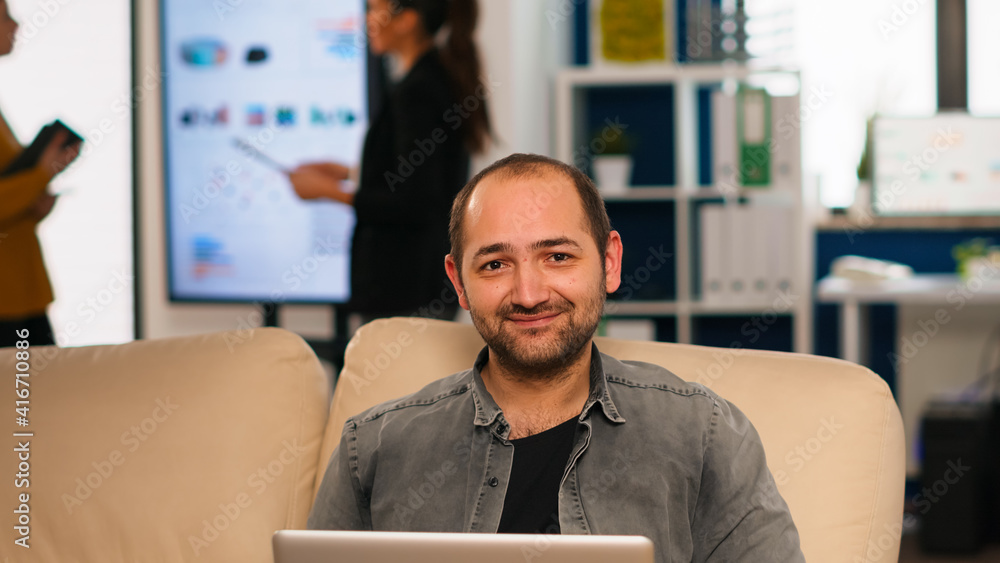 Poster manager sitting on couch in front of camera smiling while multiethnic team working in background wit