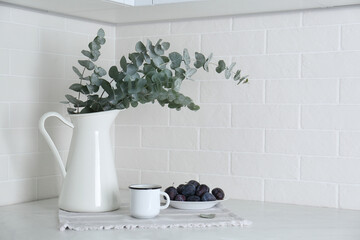 Beautiful eucalyptus branches, cup of drink and fresh plums on countertop in kitchen, space for...