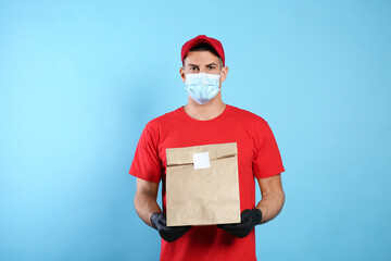 Courier in medical mask holding paper bag with takeaway food on light blue background. Delivery service during quarantine due to Covid-19 outbreak