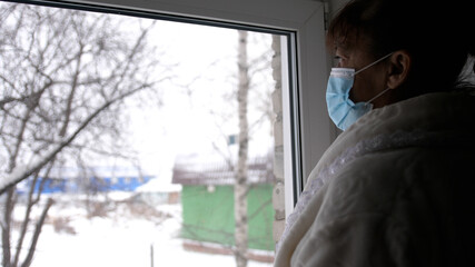 Old despair woman in protective medical mask looking sadly out window, sitting at home in quarantine. Self isolation in coronavirus pandemic. prevention of covid-19