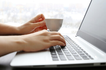 Woman drinks coffee sitting at laptop keyboard on window background. Cup of hot drink in female hand, break during work