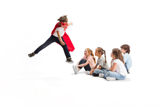 Flying. Child pretending to be a superhero with his friends sitting around him. Kids excited and inspired by their brave friend in red coat isolated on white background. Dreams, emotions concept.