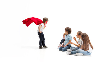 Safety. Child pretending to be a superhero with his friends sitting around him. Kids excited and inspired by their strong friend in red coat isolated on white background. Dreams, emotions concept.