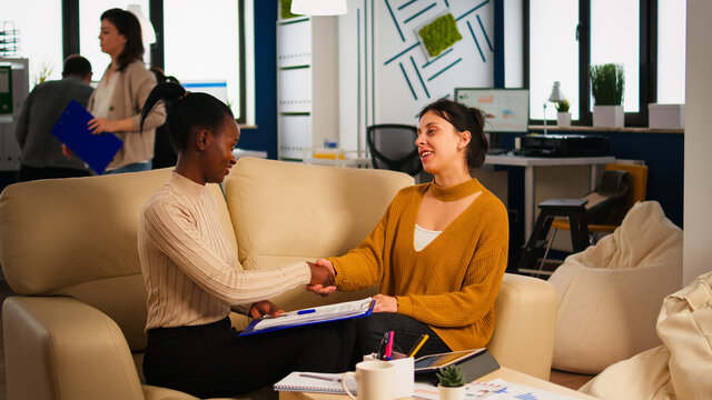 Pleased multiracial business partners sitting on couch in modern office shaking hands after signing partnership contract. Diverse team being happy for successful negotiations in start up company.