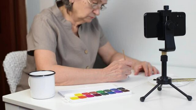 Senior woman in glasses painting flowers with watercolors, indoors. A pensioner taking an online drawing course. Selective focus on smartphone