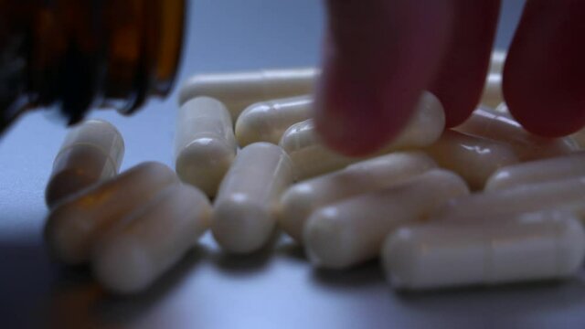Close-up, fingers take one pill. Tablets in capsules are scattered on a glossy surface, next to it lies a glass bottle for tablets. Slow refocus on pills, fingers taking one pill.
