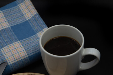 Una taza blanca con cafe, una  tostada de pan con un bollo de leche sobre un plato de madera y una mantel azul sobre fondo negro 
