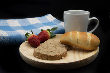 Una taza blanca con cafe, una tostada y un bollo de leche, fresas sobre un plato de madera y un mantel azul, todo sobre fondo negro.