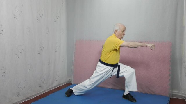 An old man athlete in a yellow T-shirt and with a blue belt trains blocks and blows with his hands in a rack