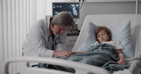 Obraz premium Aged pediatrician holding sick kid hands and sleeping at bedside