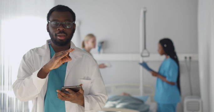 African Man Doctor Looking At Camera And Talking With Colleagues Treating Patient On Background