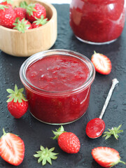 A pot of homemade strawberries jam on a black table with its fruits in scene, fruit preserving and confectionery concept   