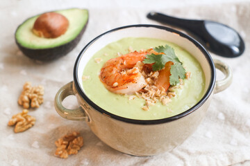 Close up of selective focus of healthy chilled avocado soup in a bowl with grilled shrimps topped with ground walnuts and coriander leaves, its fruit in background , ketogenic diet concept 