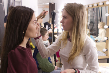 Girl makeup artist, blonde, applying makeup to a girl of 25, European appearance with long dark hair. Process, makeup artist work, beauty industry, generation y. Model preparation in the dressing room
