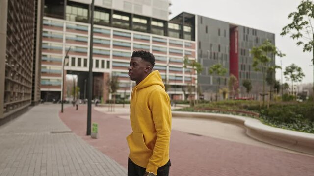 Stylish portrait of a cool black african american man in a yellow hoodie walking on modern street. He is checking time with wrist watch. Shot in 4k. City exploring