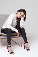 A beautiful girl of 25 years old, European appearance, with dark long hair, in a white shirt and black trousers, sits on an armchair, in a light studio, straightens her hair and stares intently