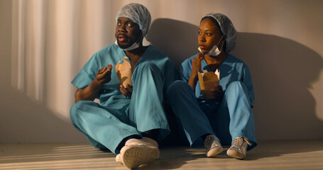 Afro-american doctors eat snack together sitting on floor in hospital corridor