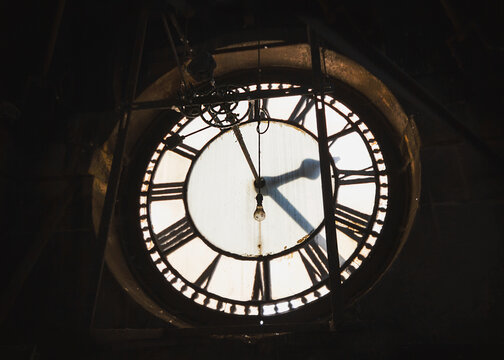 Inside abandoned old ancient clock tower with Roman numerals gears and light bulb hanging down to illuminate from behind hour minute and second hands ticking by