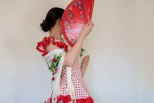 Mujer joven con un hermoso traje de flamenca de lunares rojos y un mant&oacute;n blanco. En una mano sostiene un abanico rojo que cubre su rostro. Concepto de baile t&iacute;pico de Espa&ntilde;a, sevillanas 