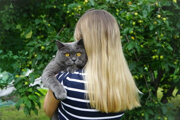 Young woman with blond  hair  gently hugs her pet grey Shorthair British cat on the in a green garden . The British Shorthair looking at the camera. Walking with pets on the street  in summer. 