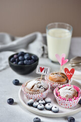 Blueberry muffins in paper tins, sprinkled with powdered sugar, with I love you flags, on a white marble board, in the background a bowl with blueberries and a glass of milk.