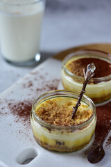 Two jars of cheesecake, sprinkled with cocoa powder, on a white marble board, with a teaspoon, a glass of milk in the back.