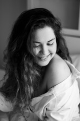 Pretty young caucasian woman on bed in modern apartment smiling after wake up. Curly hair. Girl in white shirt smiles to camera, braces teeth. Calm and cozy.