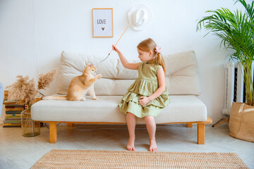 Cute little girl in a green dress playing with a cat at home on the couch in the living room